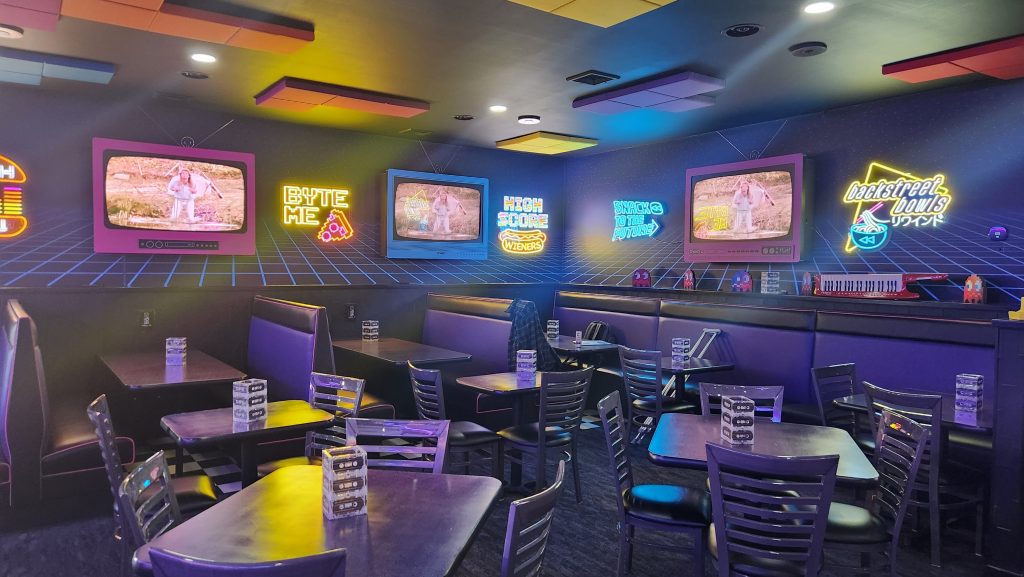 Retro arcade dining area at RWND Restaurant & Arcade with neon signs and vintage TVs.